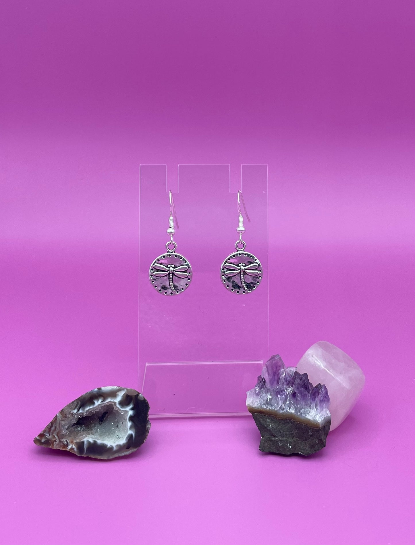 Silver circular dragonfly earrings on a clear stand with amethyst crystal on a pink background