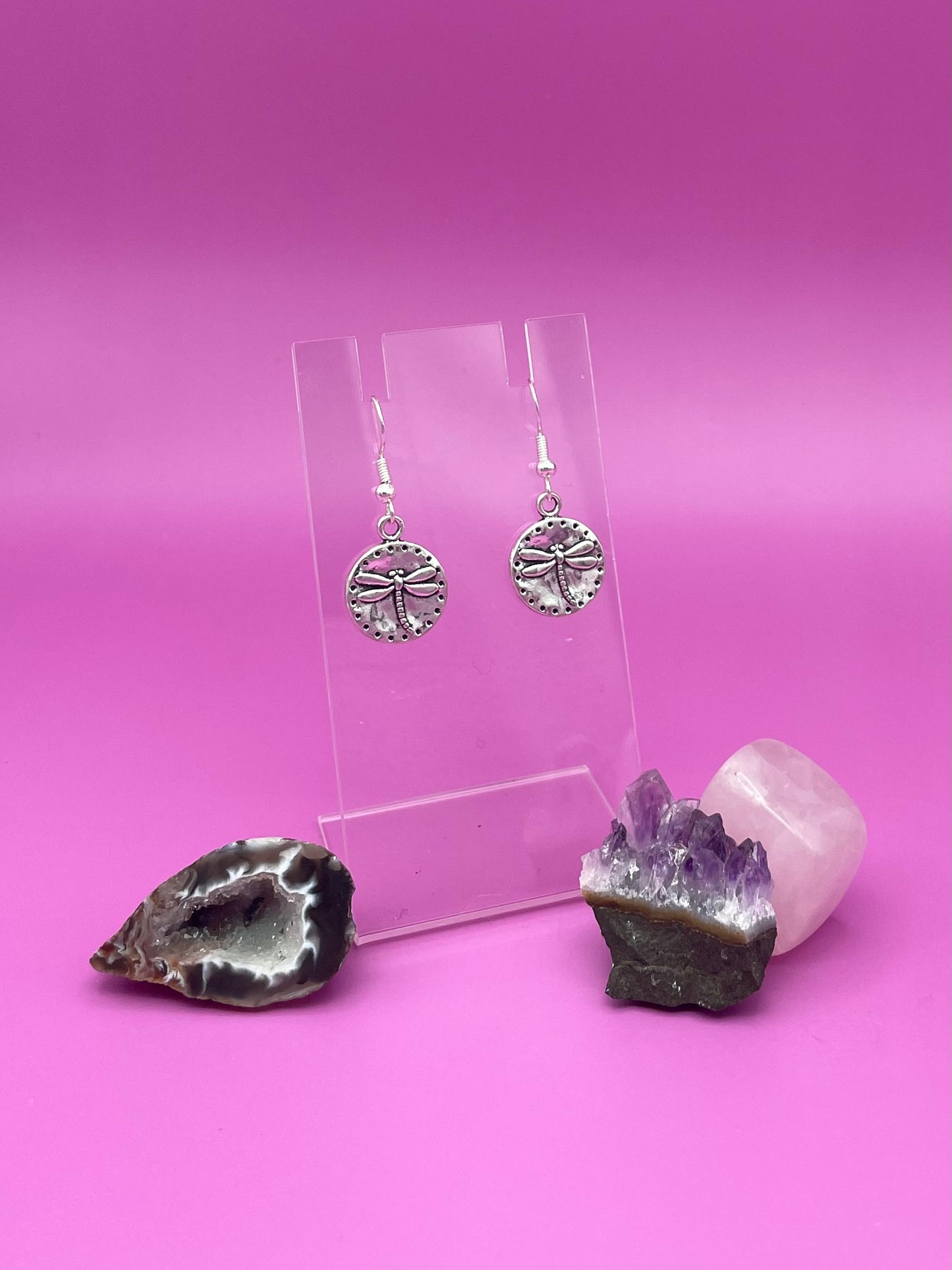 Silver circular dragonfly earrings on a clear stand with amethyst crystal on a pink background