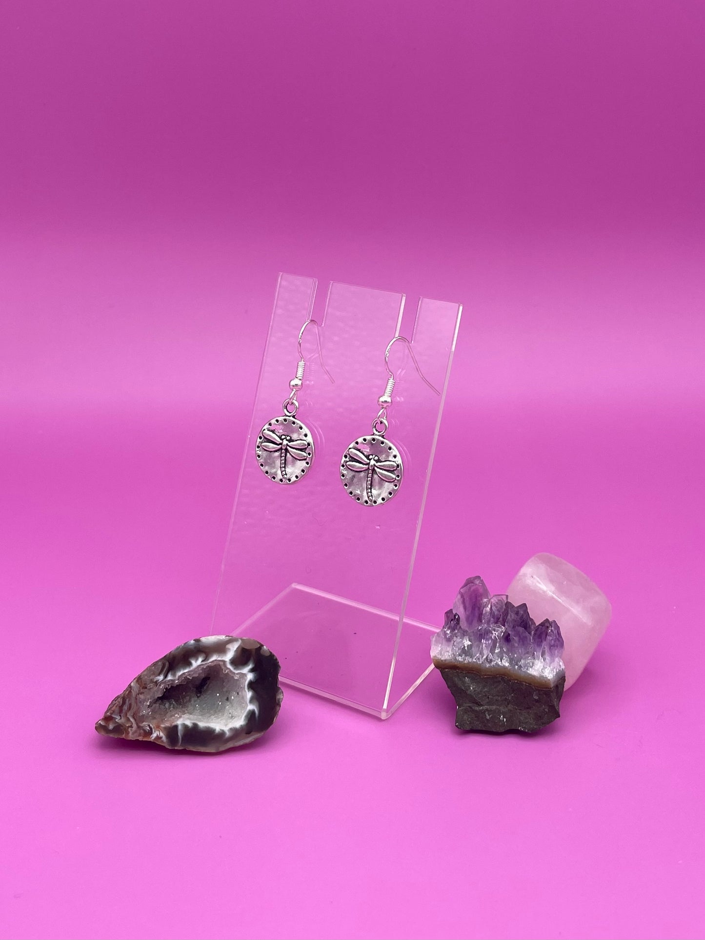 Silver circular dragonfly earrings on a clear stand with amethyst crystal on a pink background