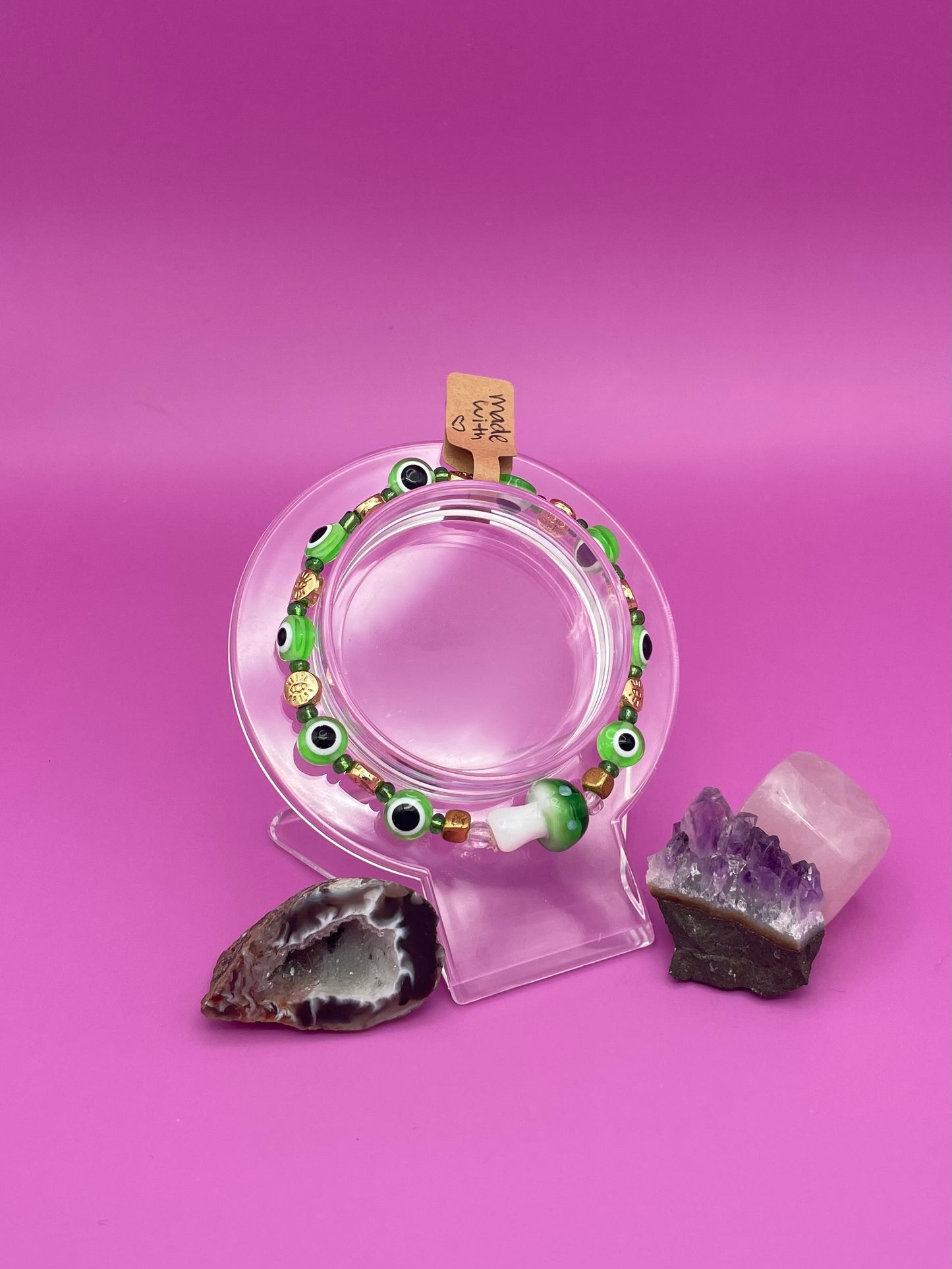 Bracelet with green evil eye beads and main glass green mushroom bead on a clear stand against a pinkish purple background