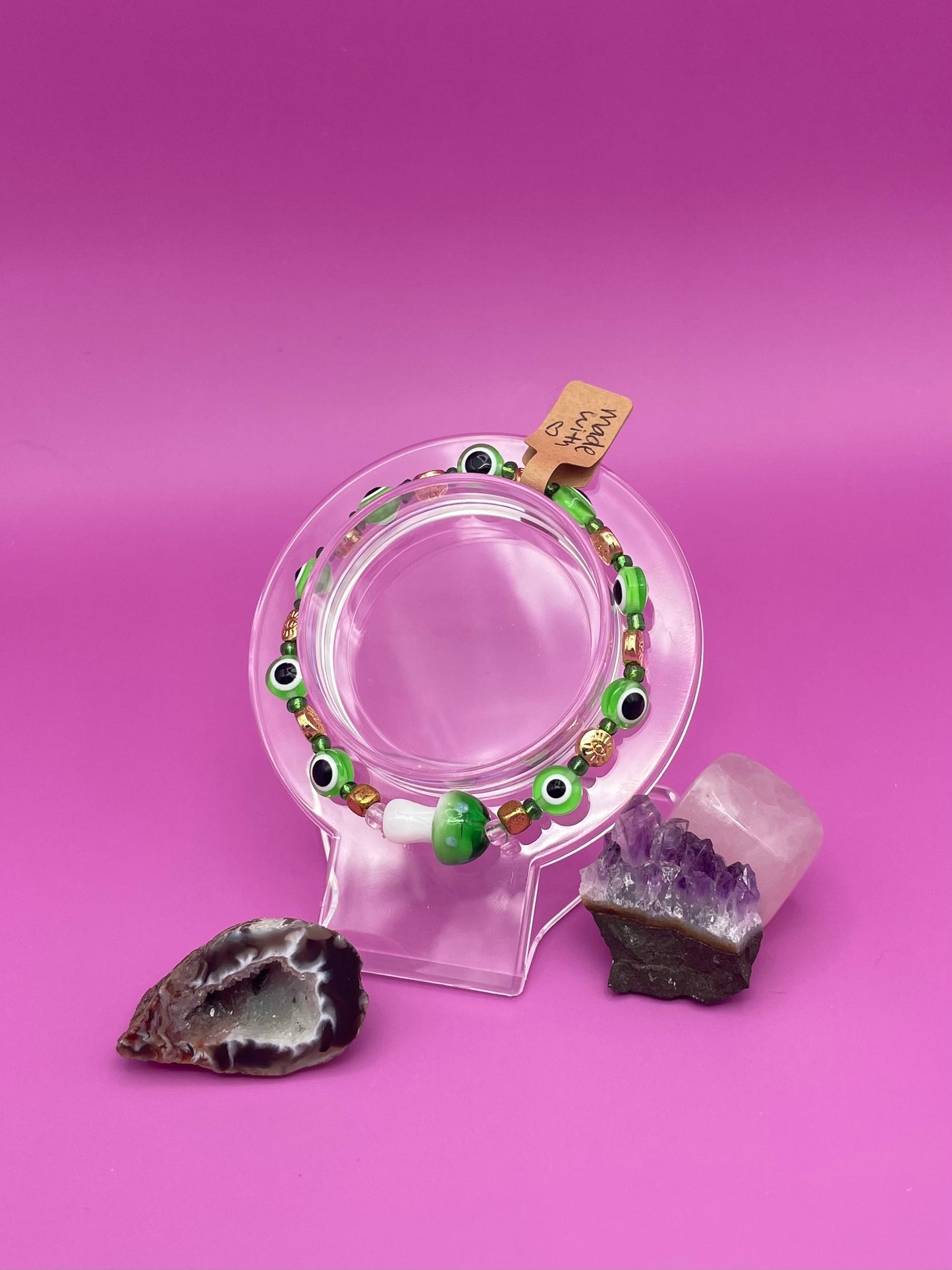 Bracelet with green evil eye beads and main glass green mushroom bead on a clear stand against a pinkish purple background