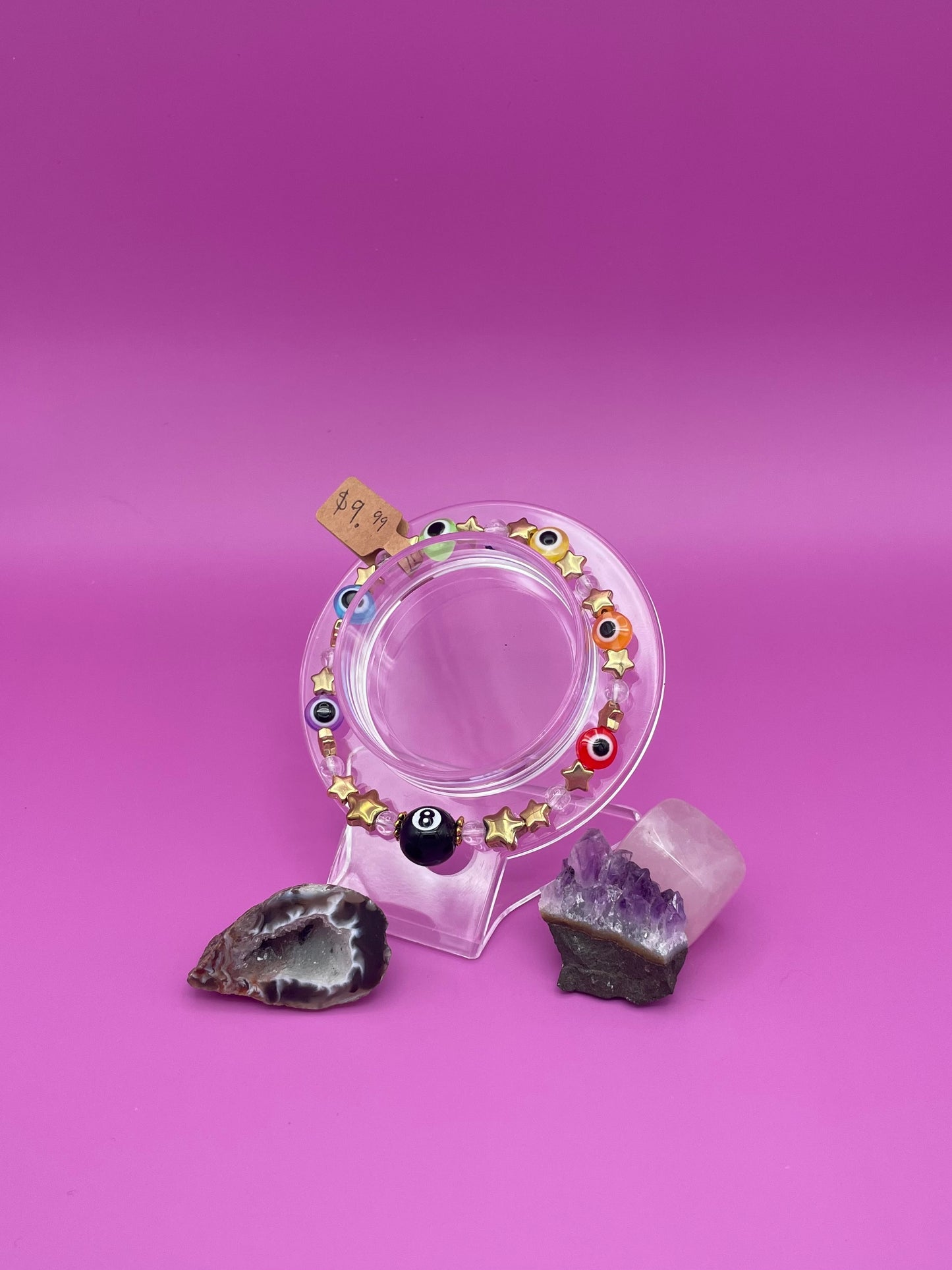 Pride evil eye bracelet with gold stars and an 8 ball charm on clear stand, surrounded by crystals on a pink background