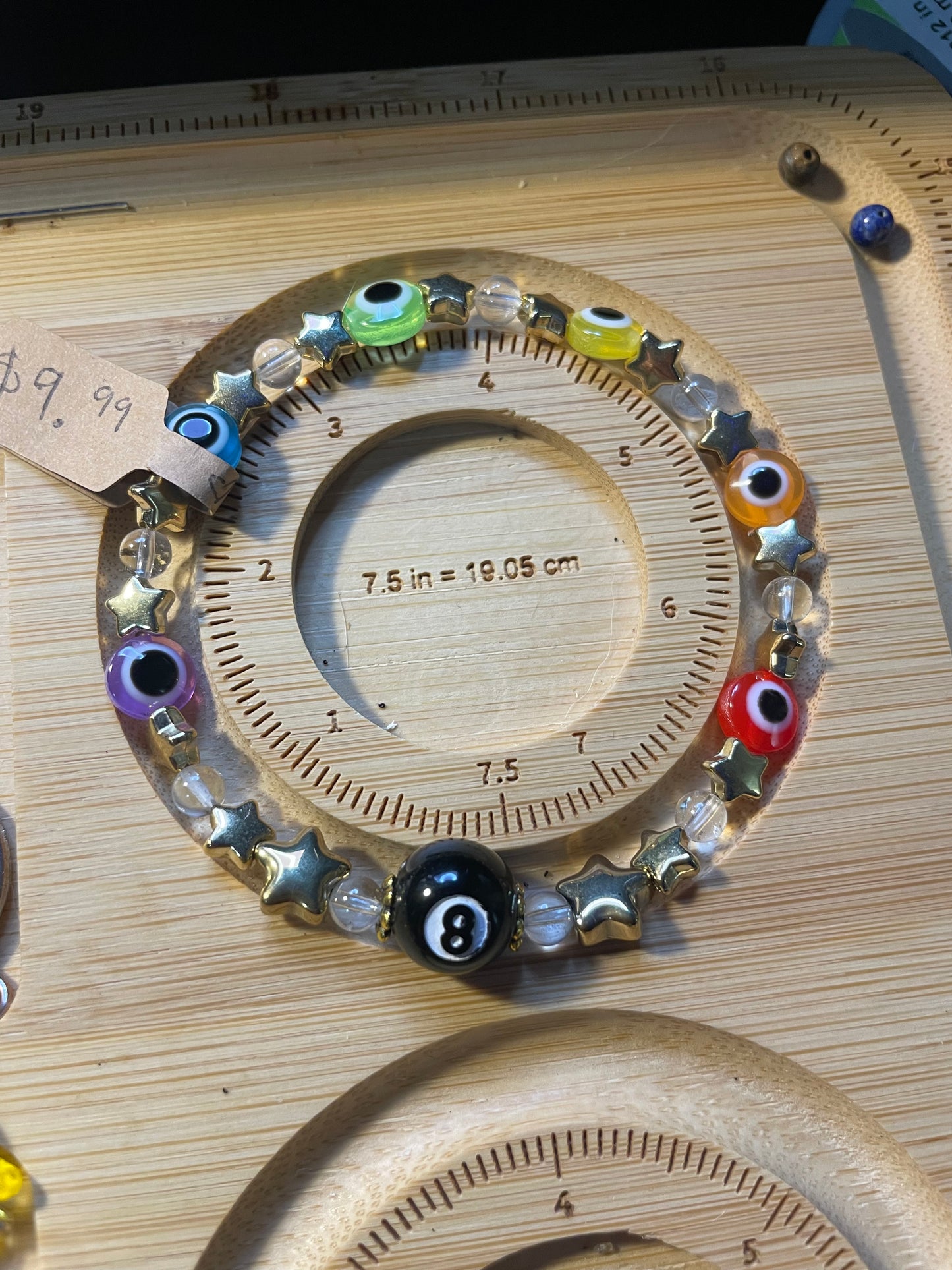 Bracelet with evil eye rainbow beads, gold stars, and clear quartz beads in a pattern with a black 8ball as the main center piece On a wooden sizing board, 7.5 inches