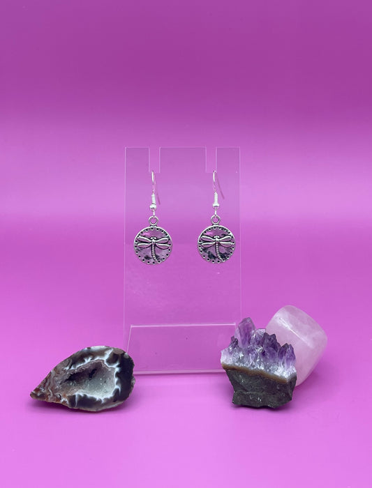 Silver circular dragonfly earrings on a clear stand with amethyst crystal on a pink background