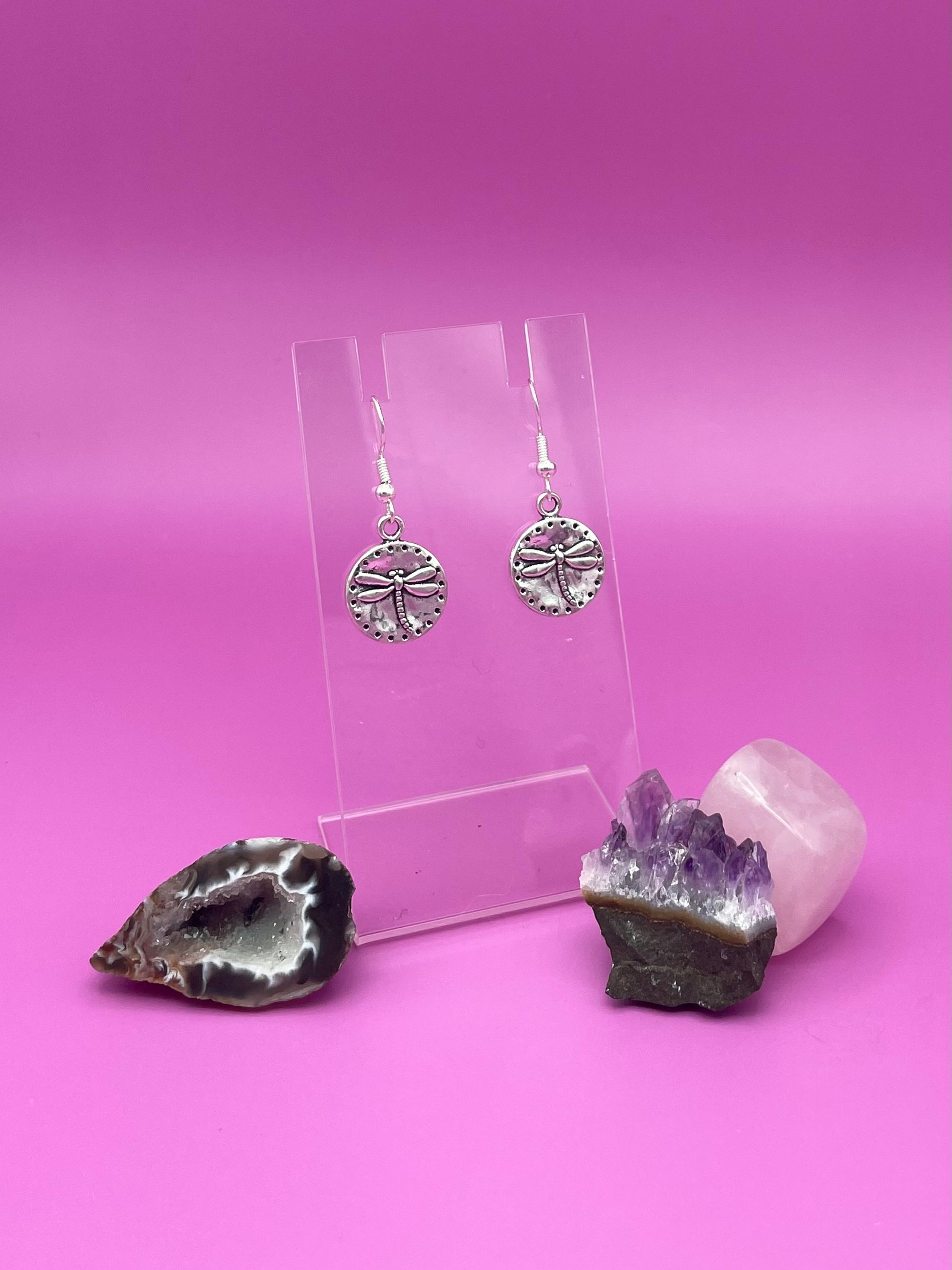 Silver circular dragonfly earrings on a clear stand with amethyst crystal on a pink background