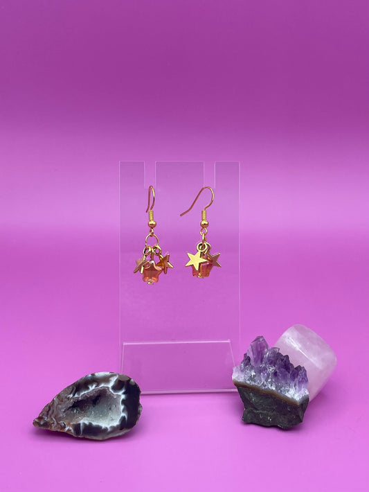 Three dangling stars earrings on a clear stand with crystals on a pink background