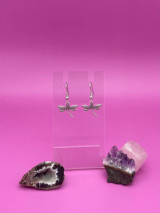 Dragonfly-shaped silver earrings on a clear stand with purple background