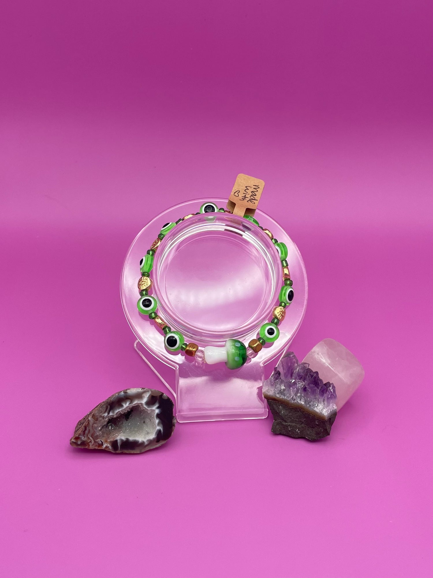 Bracelet with green evil eye beads and main glass green mushroom bead on a clear stand against a pinkish purple background