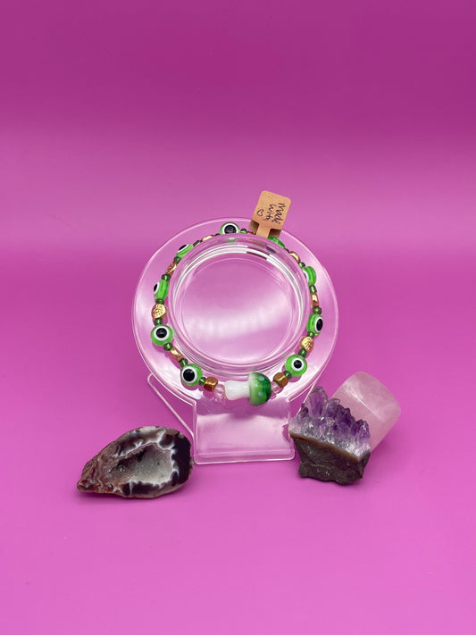 Bracelet with green evil eye beads and main glass green mushroom bead on a clear stand against a pinkish purple background